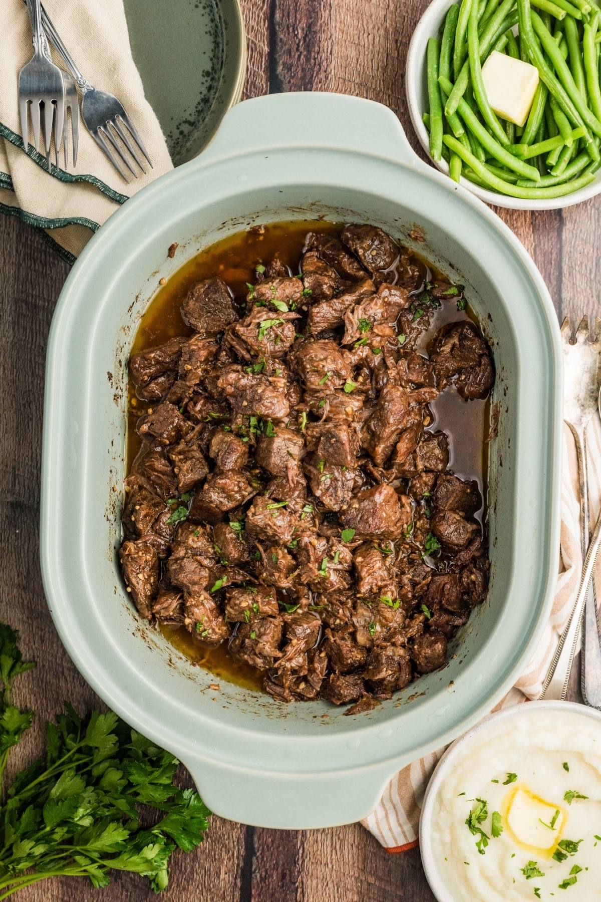 Beef tips in sauce in a slow cooker, with mashed potatoes and green beans on the side.