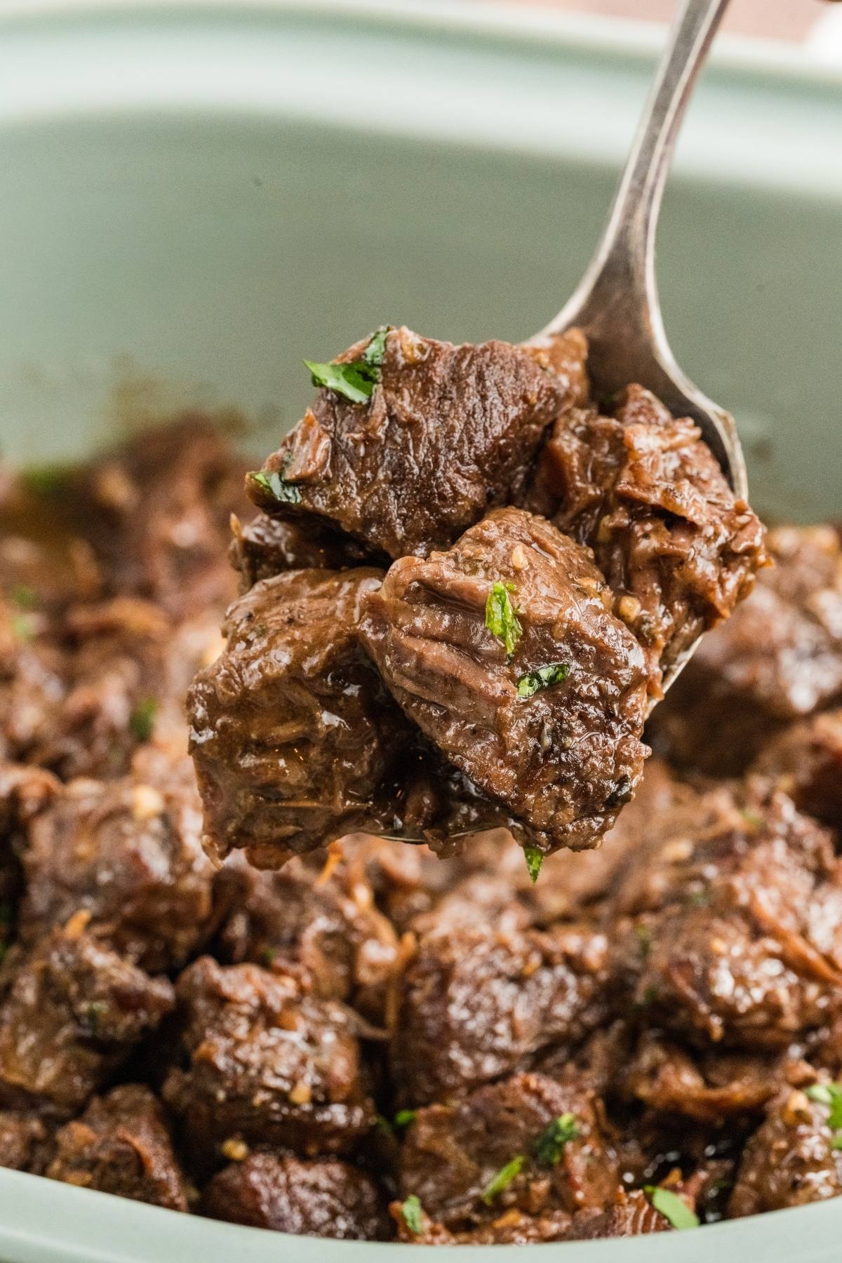 A spoon lifts tender, cooked beef chunks garnished with herbs from a bowl of braised meat.
