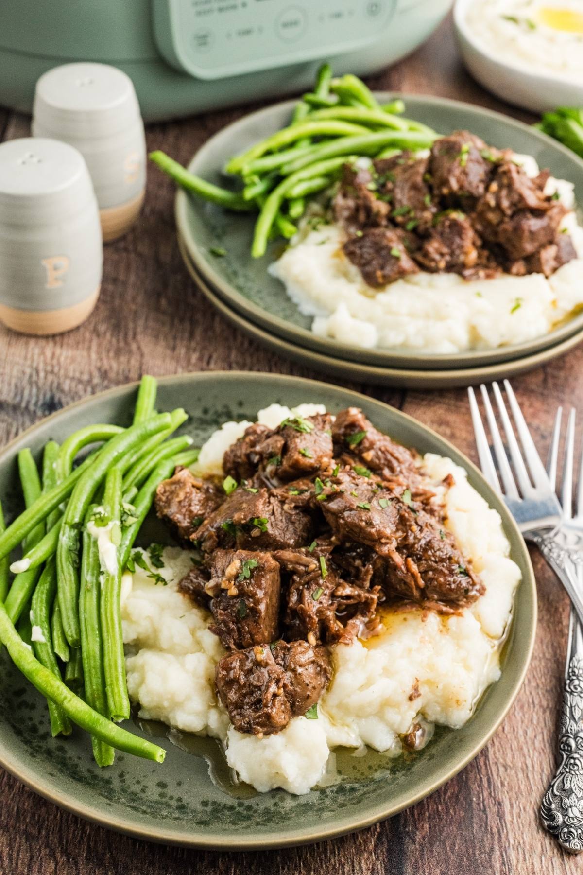 Plates of slow cooker steak bites over mashed potatoes with green beans on the side, served on a wooden table.