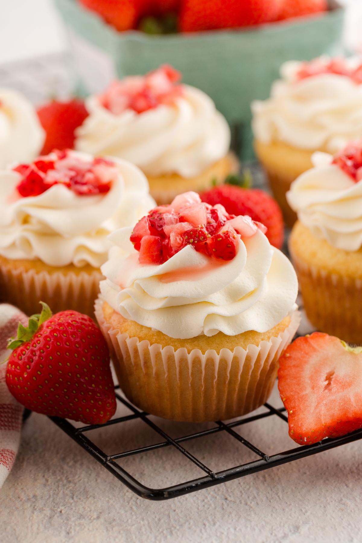 Vanilla cupcakes with white frosting and chopped strawberries on top, placed on a cooling rack with fresh strawberries.