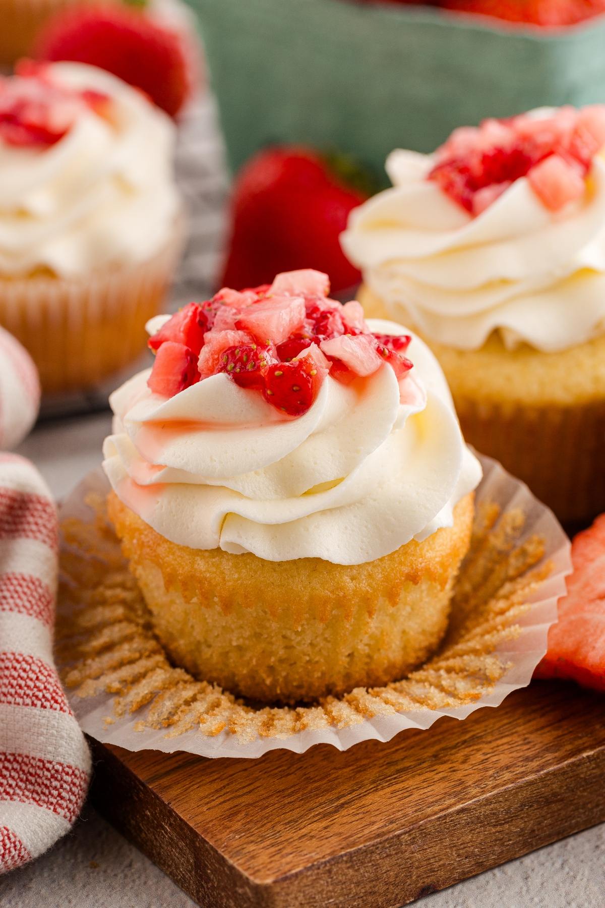 strawberry shortcake cupcakes with white frosting and chopped strawberries on top, resting on a wooden board.