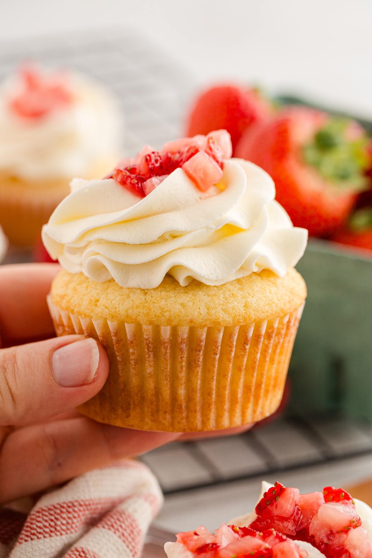 A hand holds a vanilla cupcake topped with white frosting and chopped strawberries, with more cupcakes and strawberries behind.