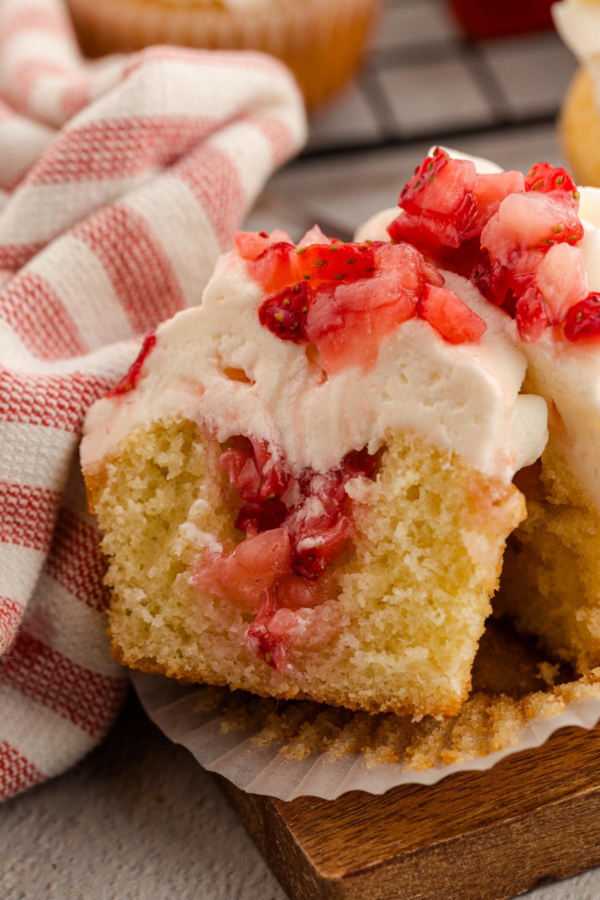 A sliced cupcake with white frosting and chopped strawberries on top, set on a wooden board near a striped cloth.