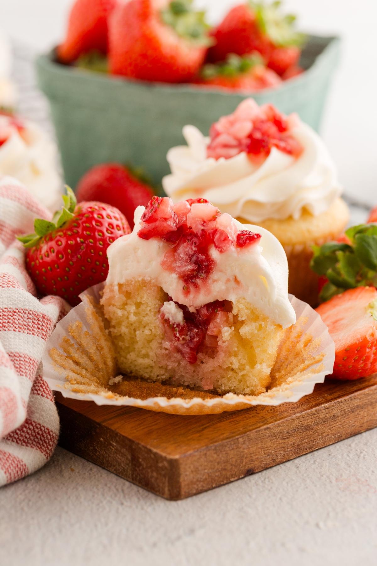 A strawberry shortcake cupcakes with a bite taken out, topped with whipped cream and strawberry pieces, surrounded by fresh strawberries.