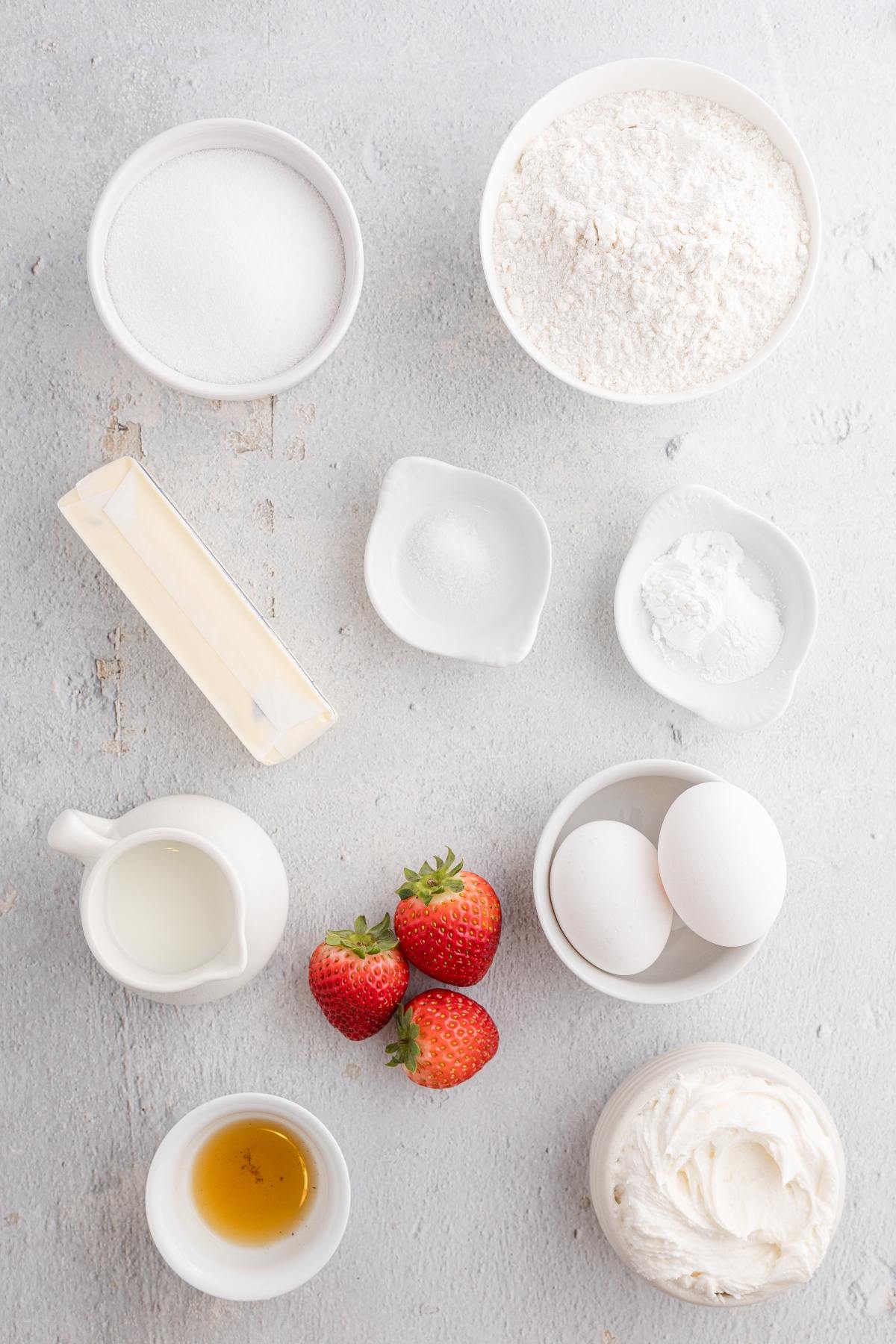 Baking ingredients arranged on a light surface, including flour, sugar, eggs, strawberries, and butter.