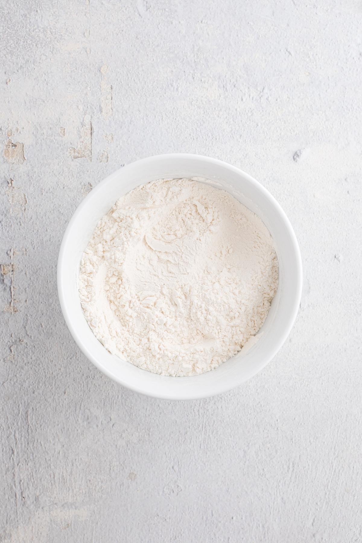 White ceramic bowl filled with flour on a light, textured surface viewed from above.