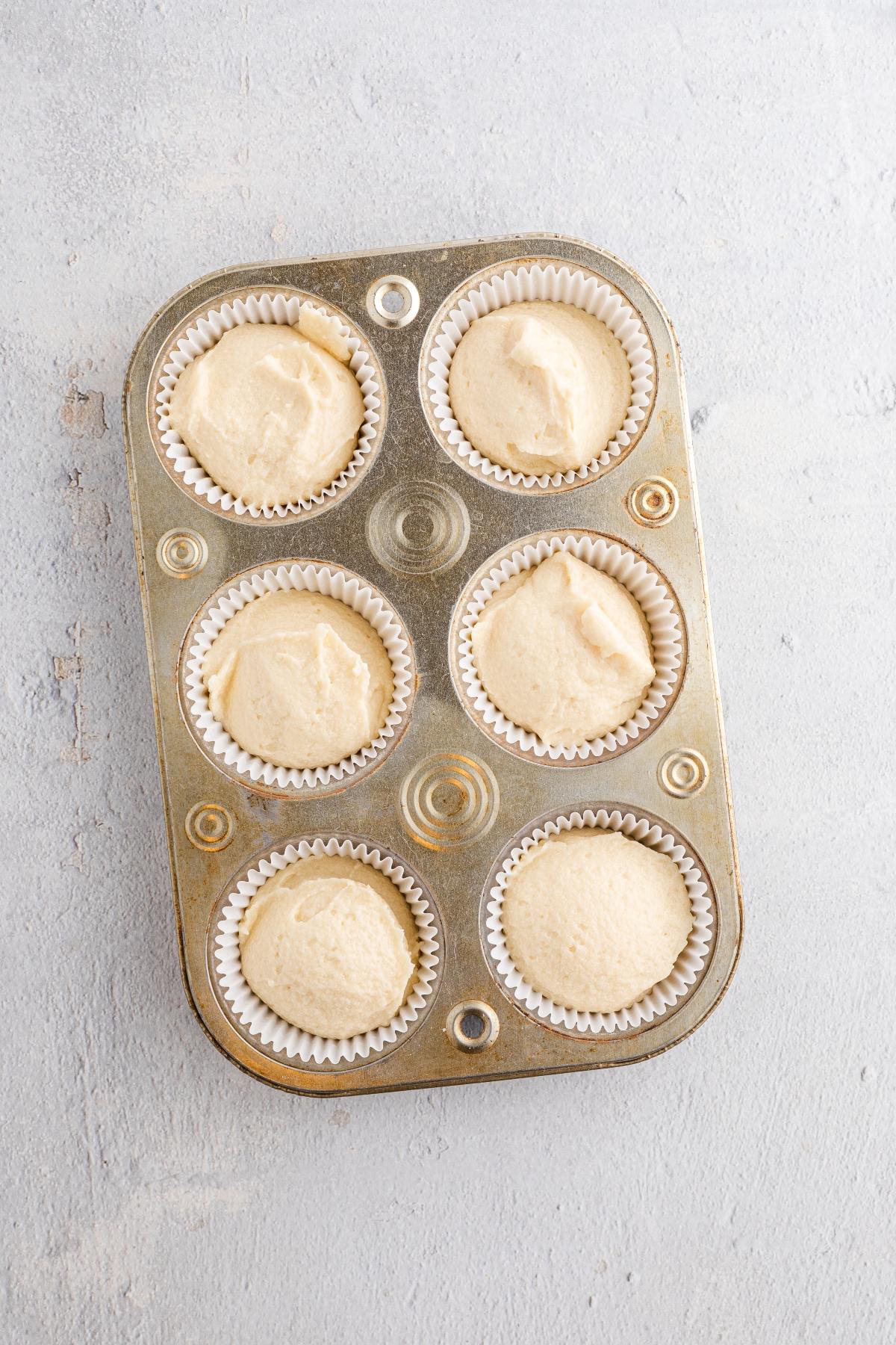 A muffin tin with six paper liners filled with raw cupcake batter on a light gray surface.