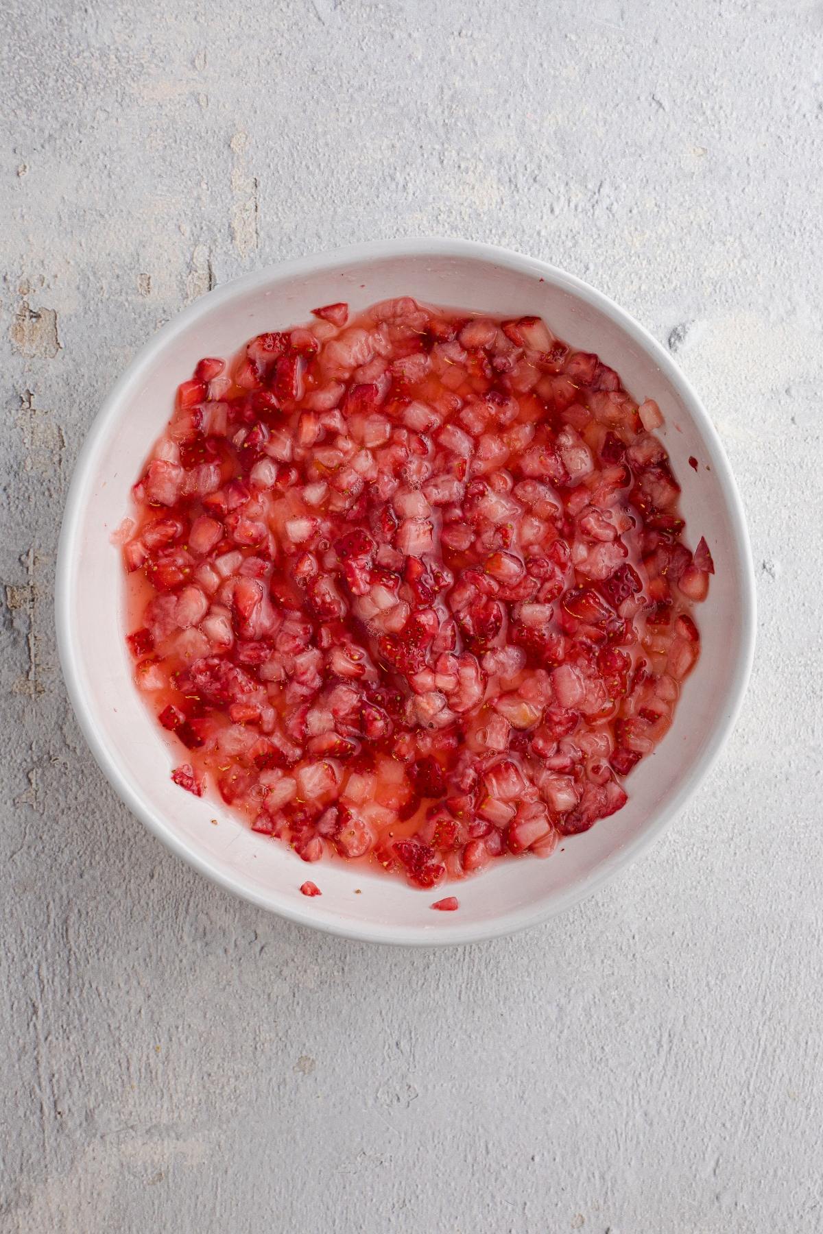 A white bowl filled with finely chopped strawberries and sugar on a light gray surface.