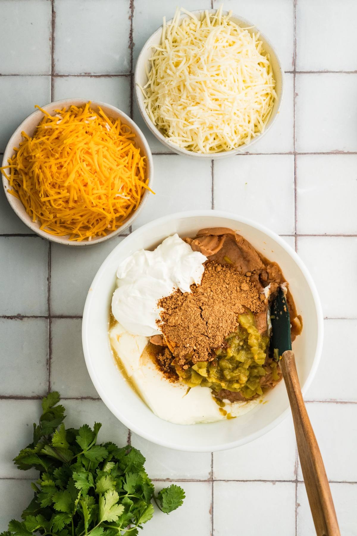 A bowl with sour cream, refried beans, cheese, spices, and chiles, surrounded by shredded cheese and cilantro.