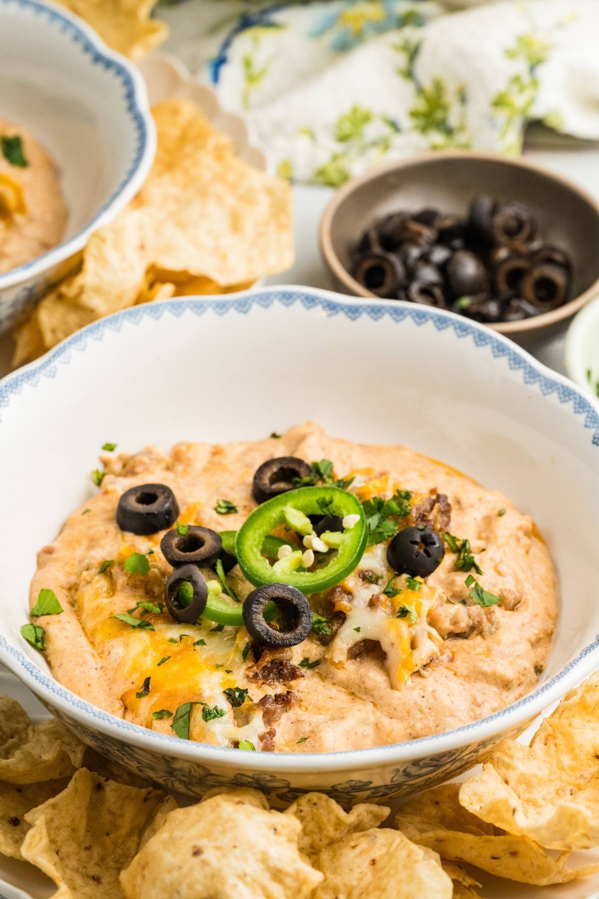 A bowl of cheesy dip topped with sliced black olives, jalapeños, and herbs, surrounded by tortilla chips.