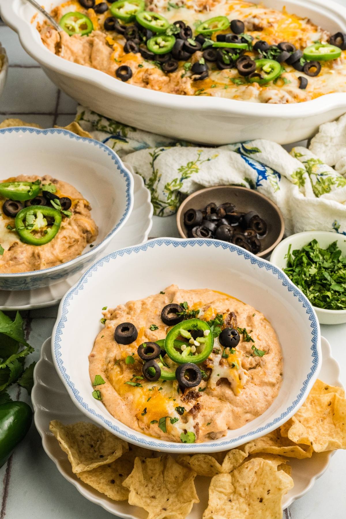 Creamy dip topped with jalapeños, olives, and herbs in bowls, served with tortilla chips on the side.
