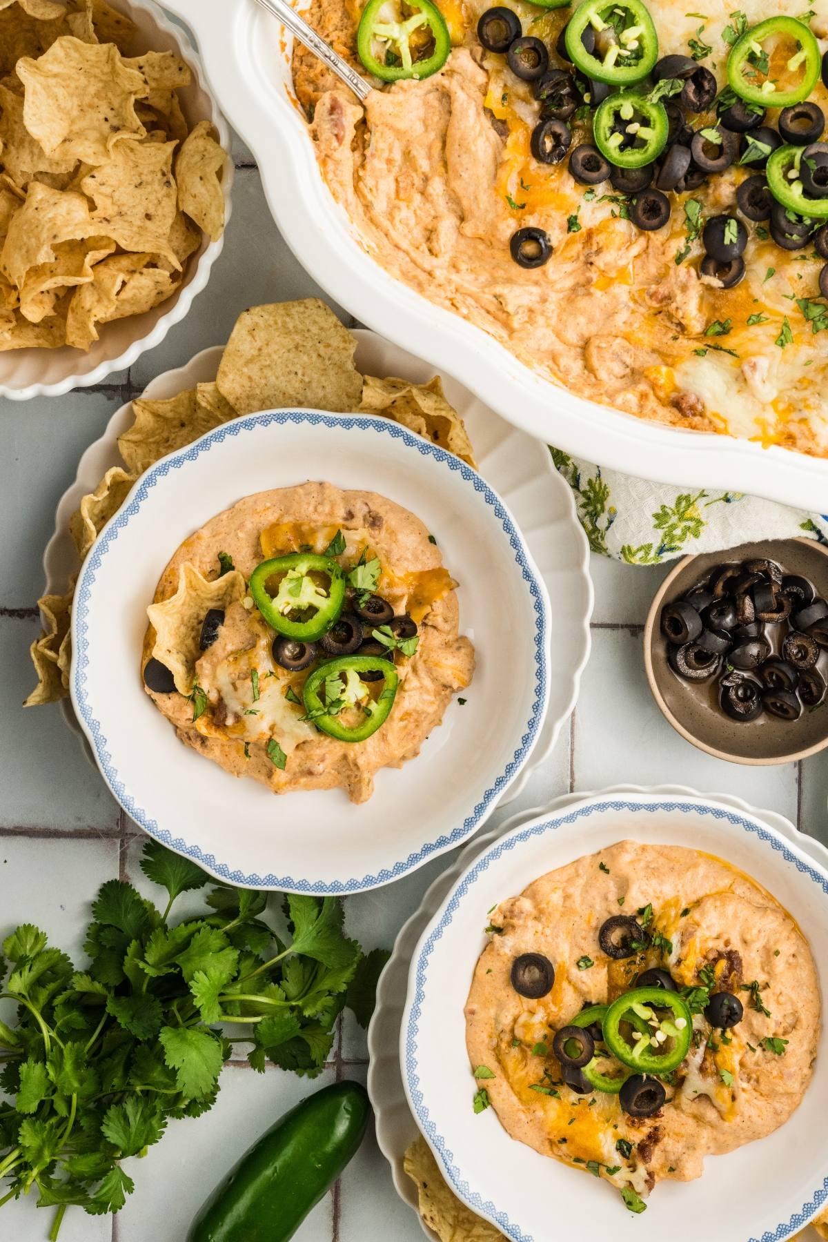 Baked cheesy dip with jalapeños and olives, served in bowls with tortilla chips, herbs, and extra olives on the side.