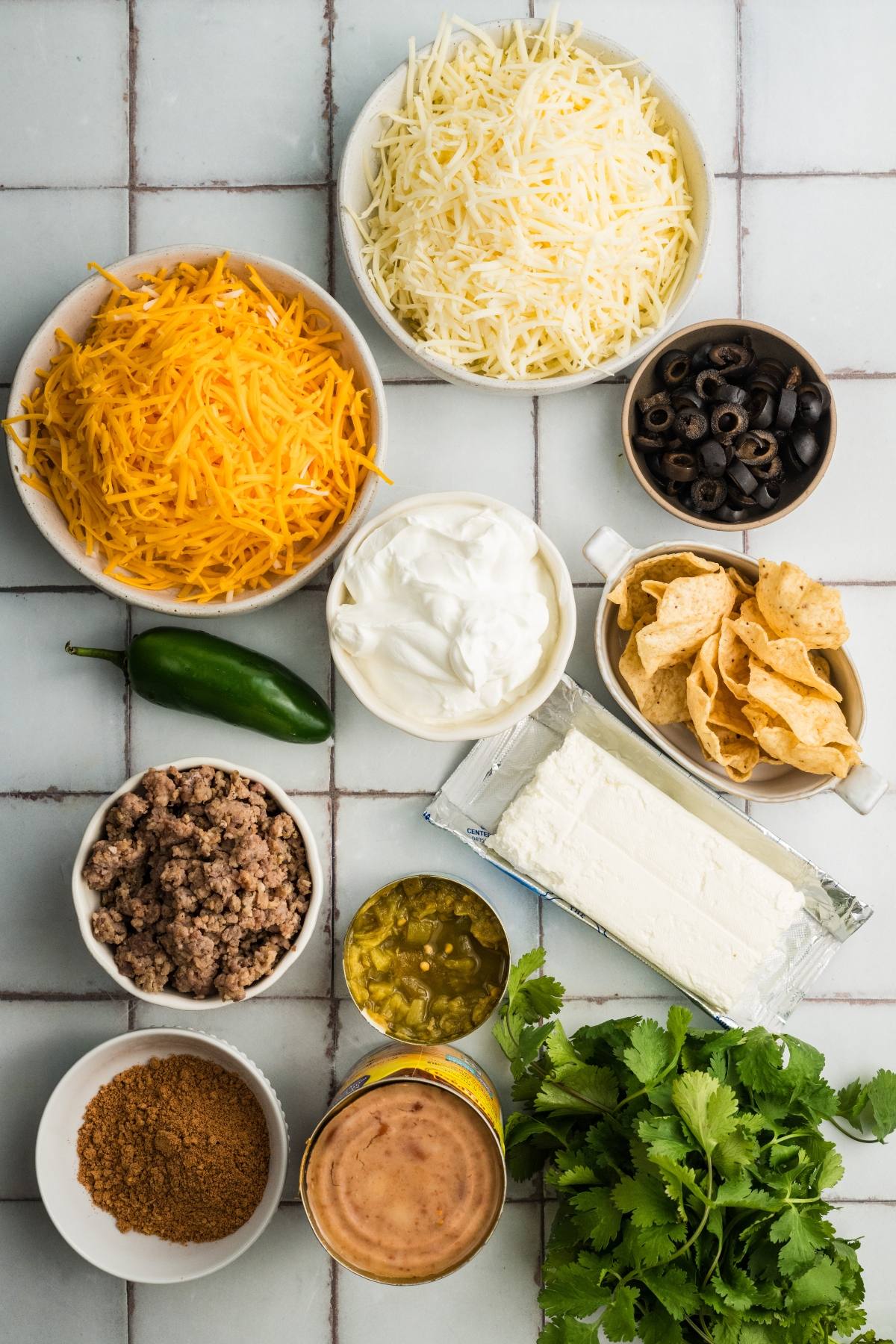 Ingredients for a dip, including cheese, sour cream, ground meat, chips, cream cheese, beans, olives, and cilantro.