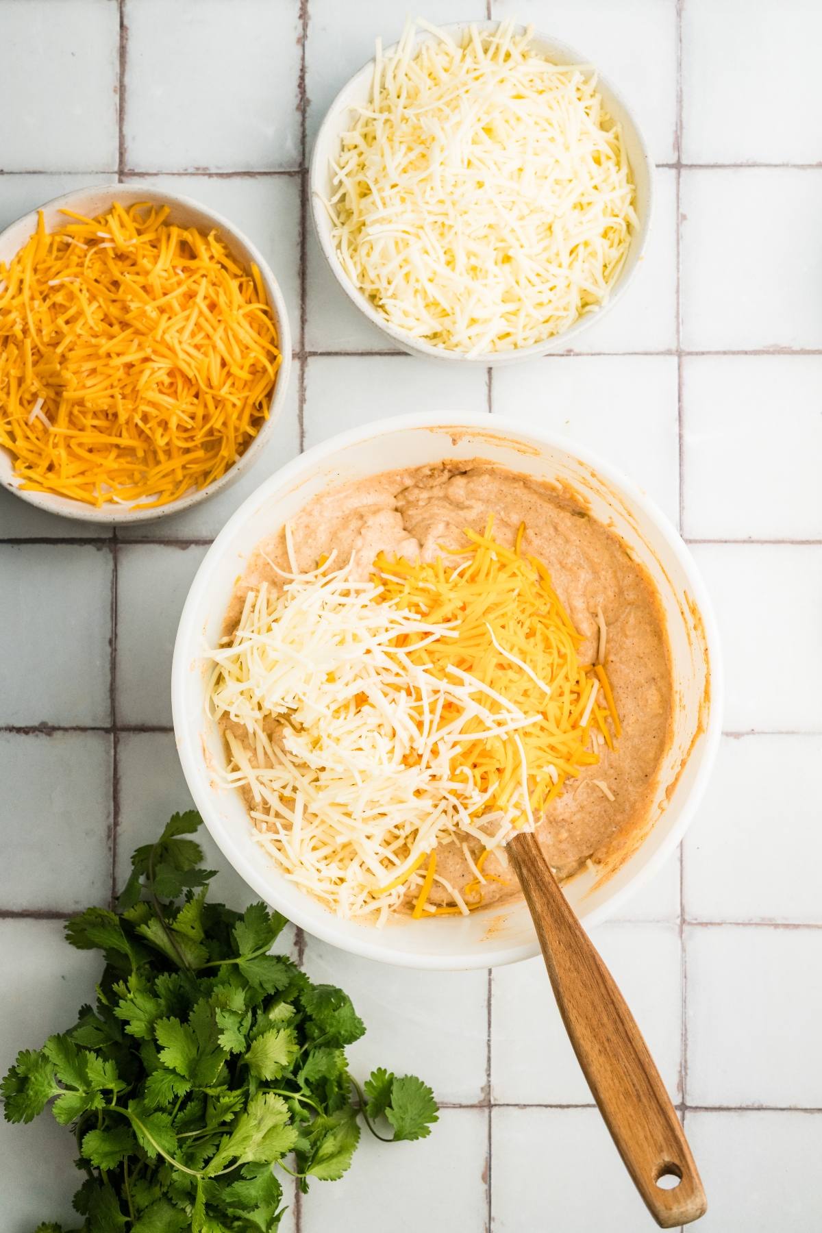 A bowl of dip being mixed with shredded cheese, surrounded by more cheese and fresh cilantro.