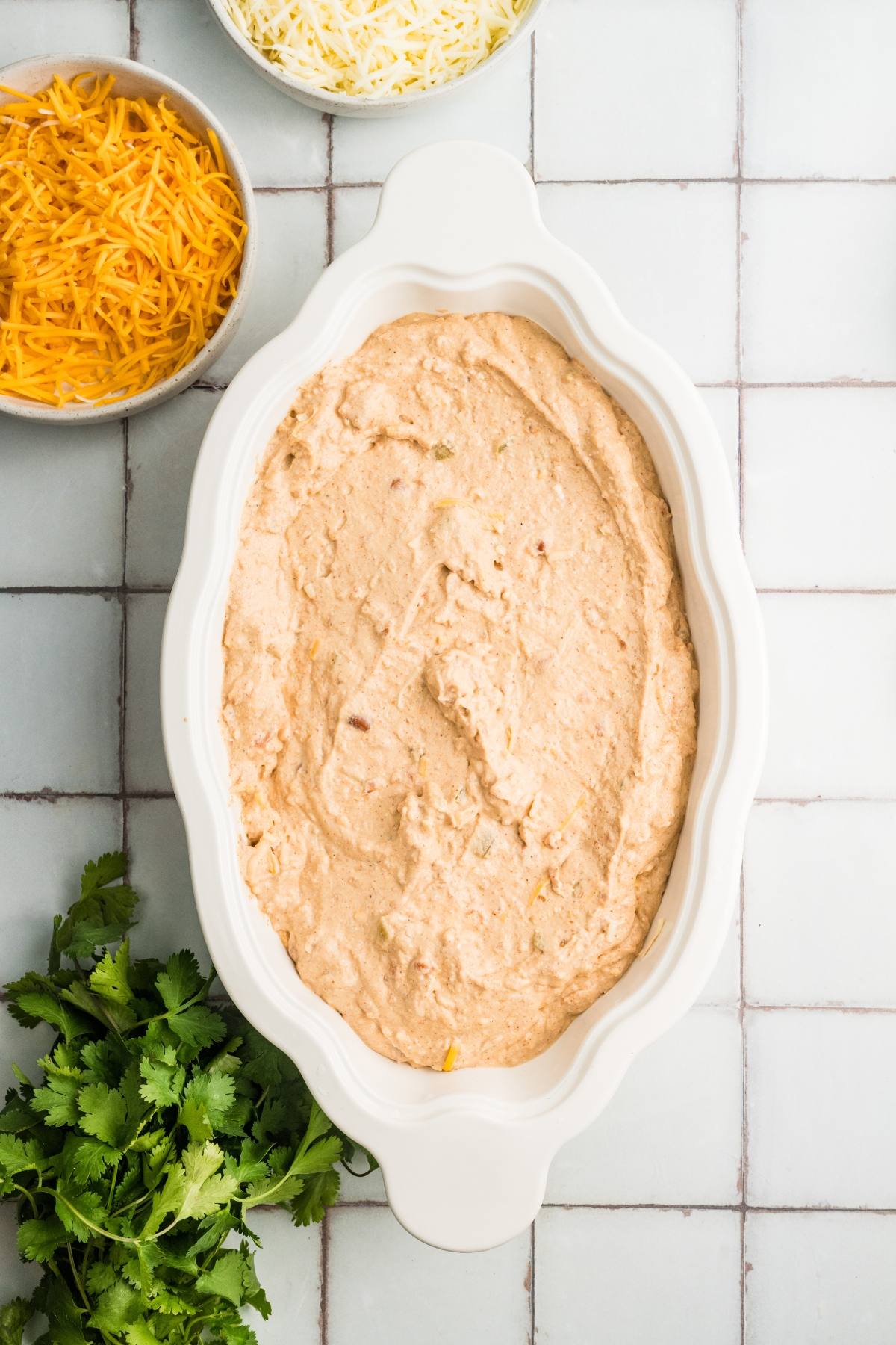 Oval dish with creamy dip mixture, shredded cheese bowls, and fresh cilantro on a tiled surface.