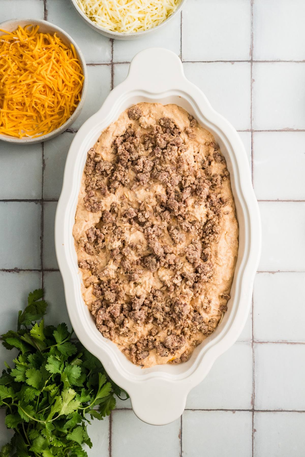 Oval casserole dish with ground beef mixture, next to shredded cheese bowls and fresh cilantro on a tiled surface.