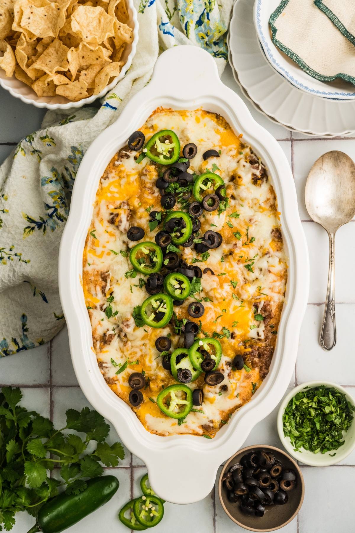 Baked casserole topped with melted cheese, sliced jalapeños, and black olives, with chips and toppings nearby.