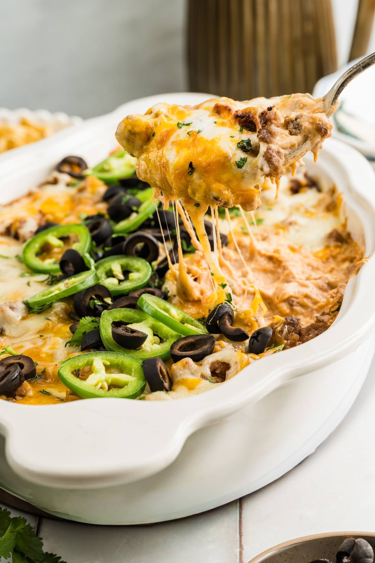 A cheesy casserole topped with sliced jalapeños and black olives, with a serving being lifted out.