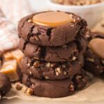 Stacked turtle cookies with caramel centers and pecan pieces rest on parchment paper, with caramels in the background.