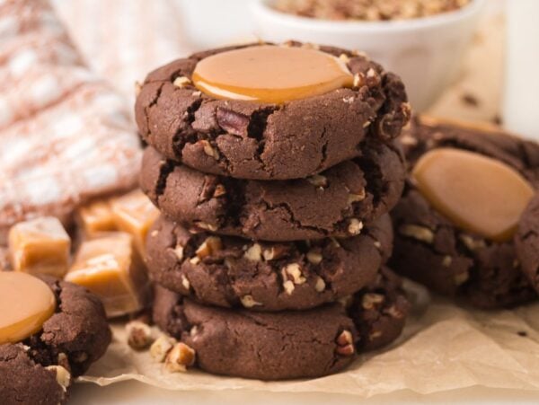 Stacked turtle cookies with caramel centers and pecan pieces rest on parchment paper, with caramels in the background.