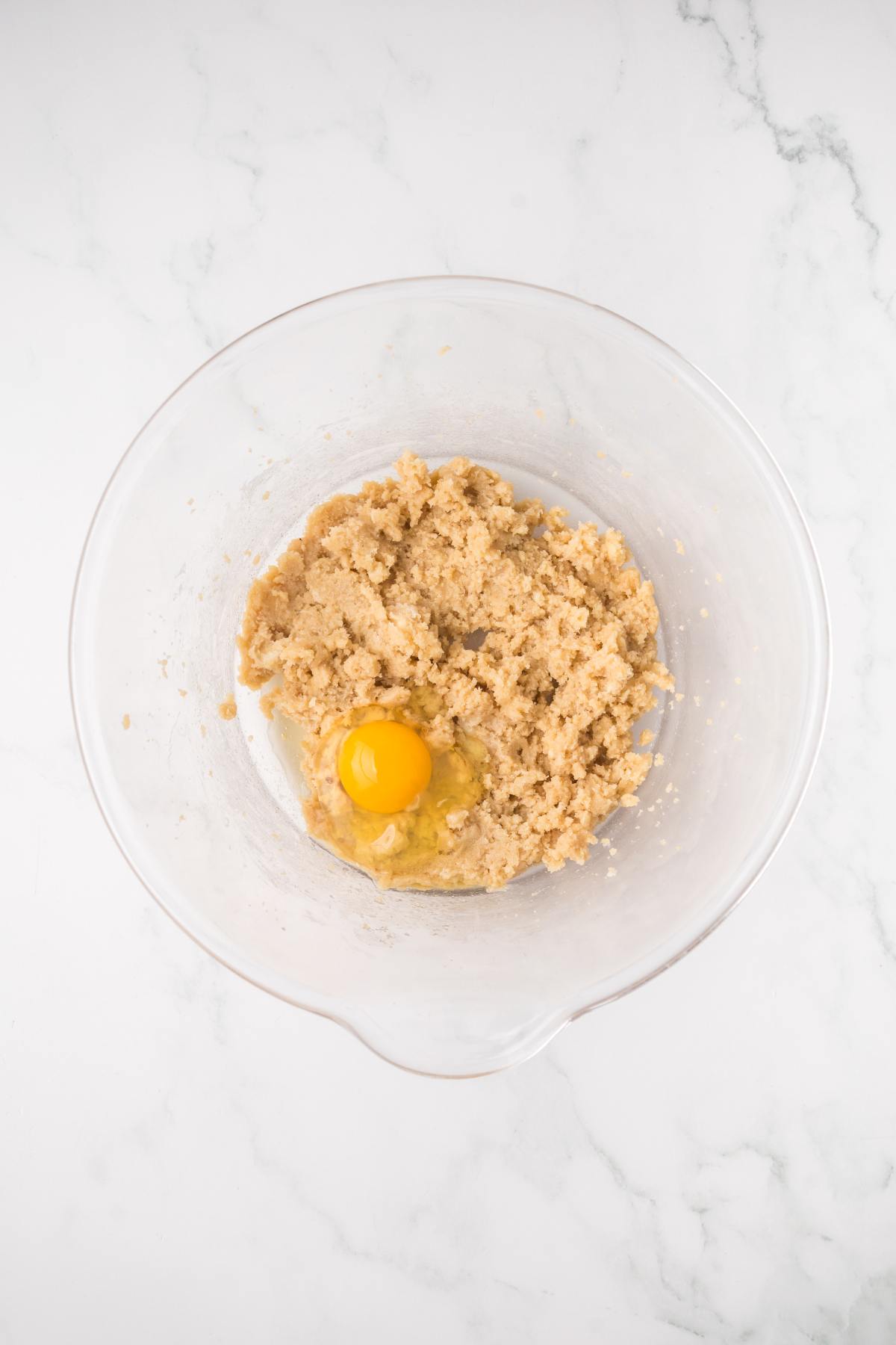 A glass bowl with creamed sugar and butter mixture and an egg on a white marble surface.
