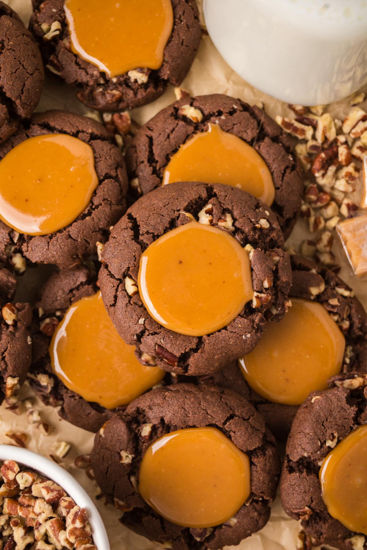 Chocolate turtle cookies with caramel centers, surrounded by chopped pecans and a glass of milk.