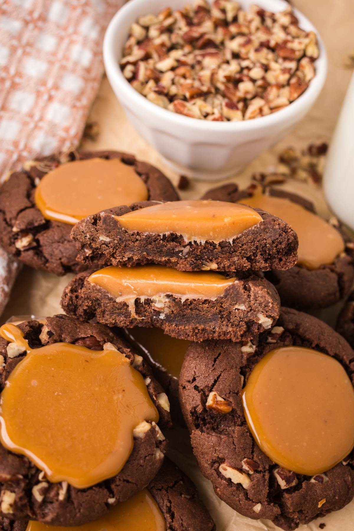Chocolate cookies topped with caramel and nuts, with a bowl of chopped pecans and a glass of milk nearby.
