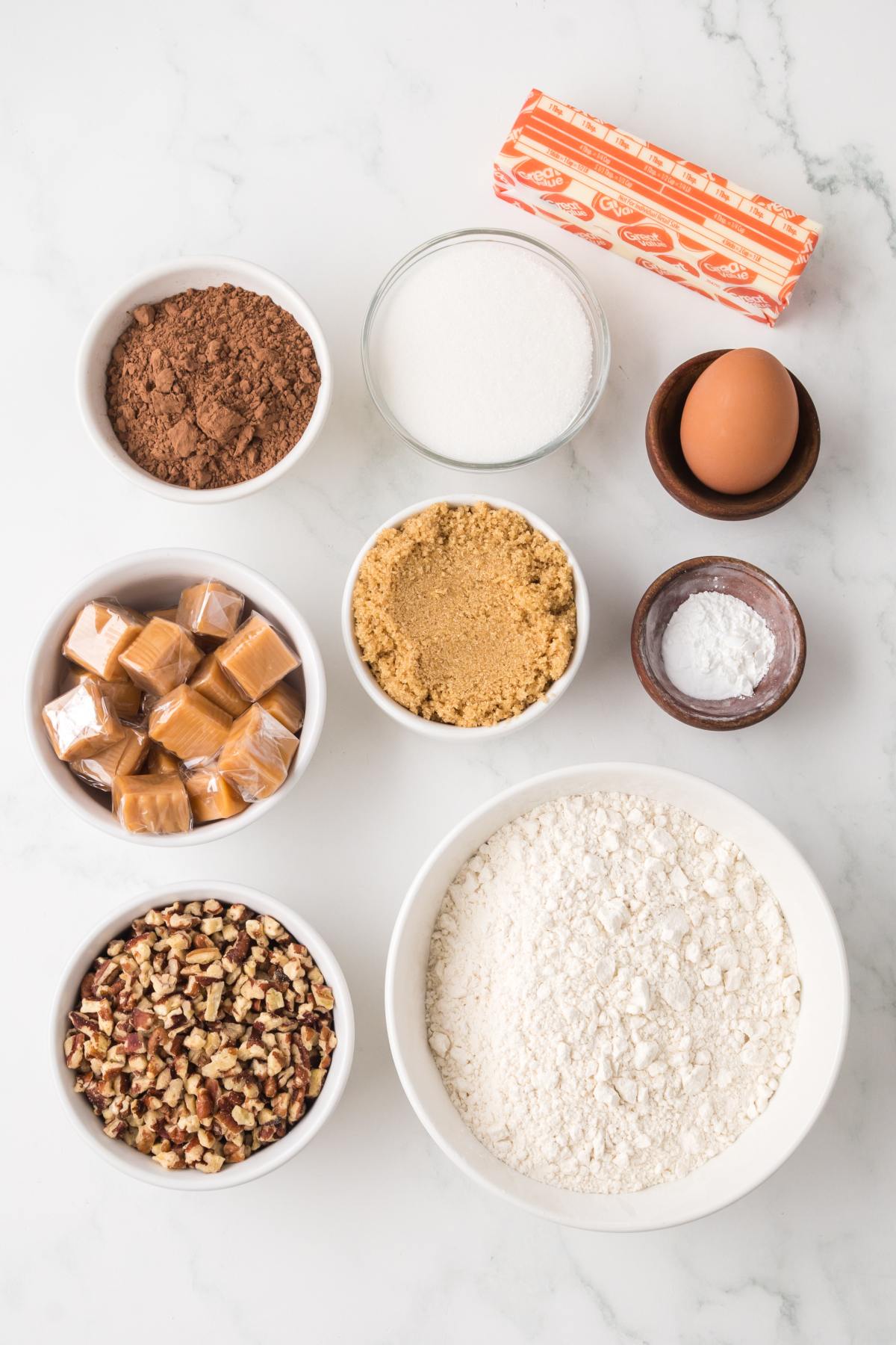 Bowls of cocoa, sugar, brown sugar, flour, pecans, egg, baking soda, butter, and caramel squares on a marble surface.