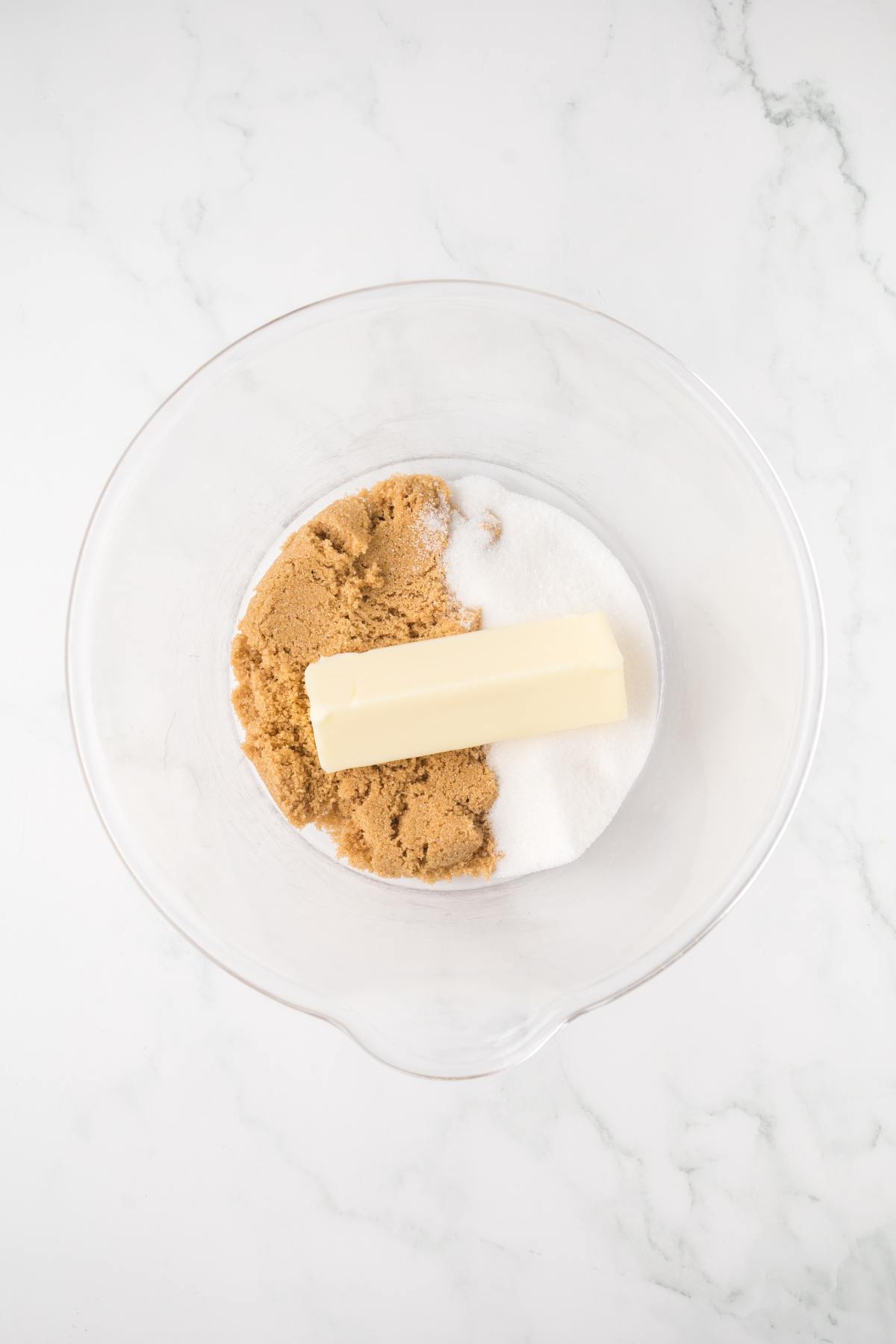 A glass bowl with brown sugar, white sugar, and a stick of butter on a white marble surface.