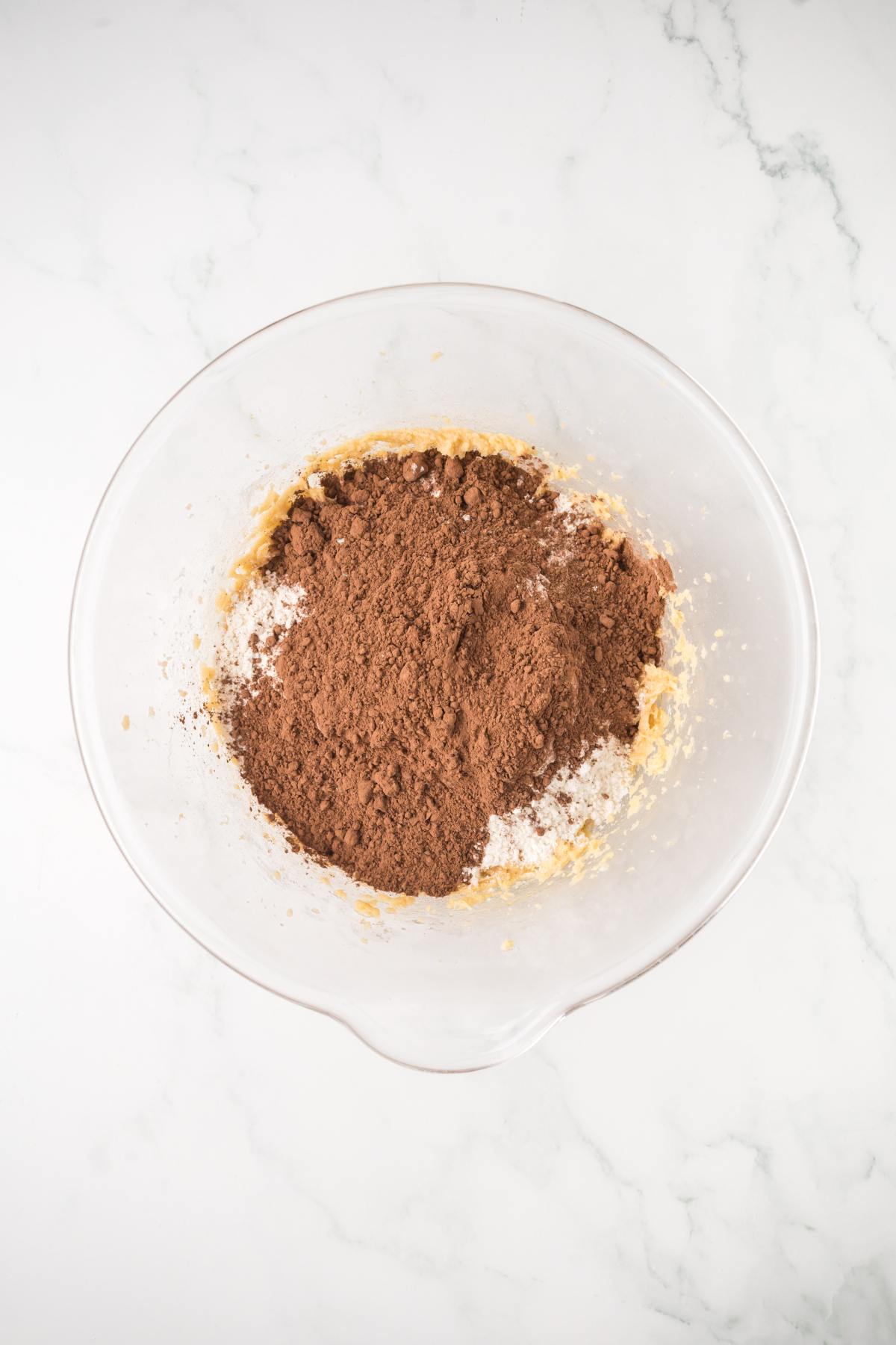 A glass bowl with flour and cocoa powder on top of a creamy mixture, on a white marble surface.