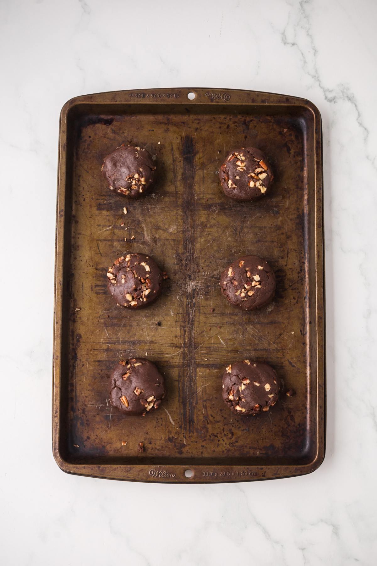 Six unbaked chocolate cookies with nuts on a large baking sheet, placed on a white marble surface.