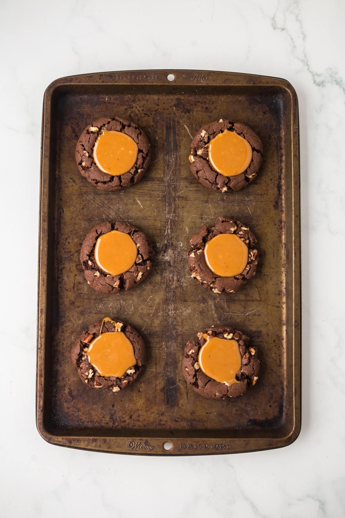 Six chocolate cookies with caramel centers on a worn baking sheet, viewed from above on a white surface.