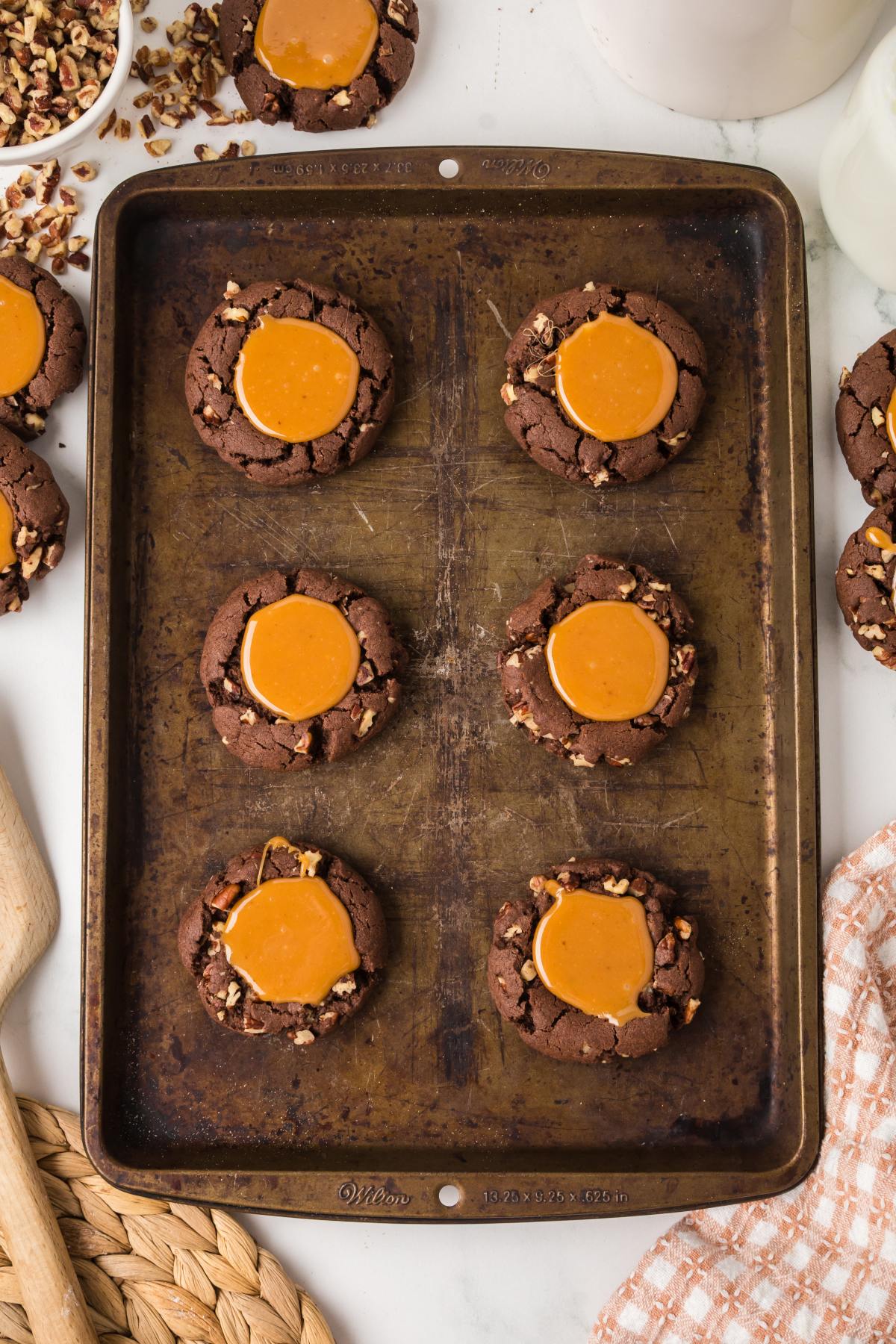 A baking sheet with six chocolate turtle cookies filled with caramel and topped with chopped nuts.