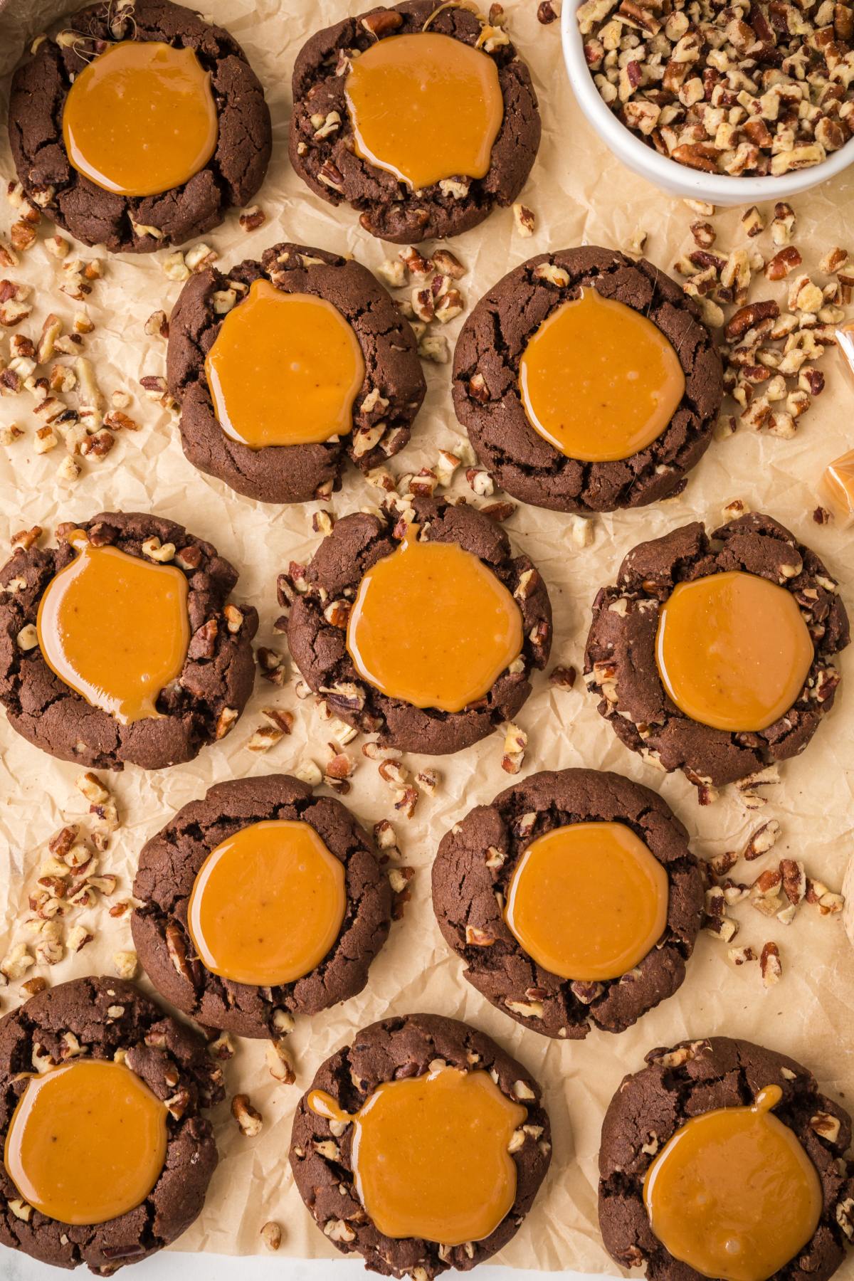 Chocolate turtle cookies with caramel centers and chopped pecans on parchment paper, with a bowl of pecans nearby.