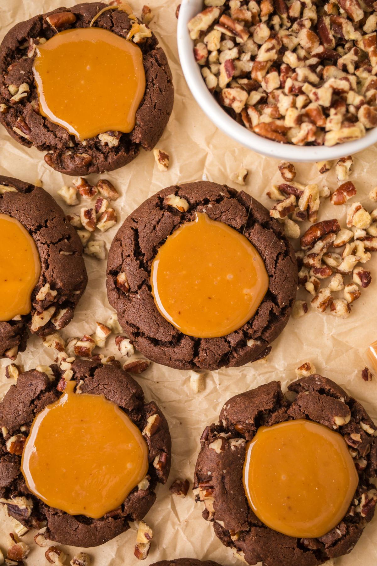 Chocolate turtle cookies topped with caramel and surrounded by chopped pecans, displayed on parchment paper.