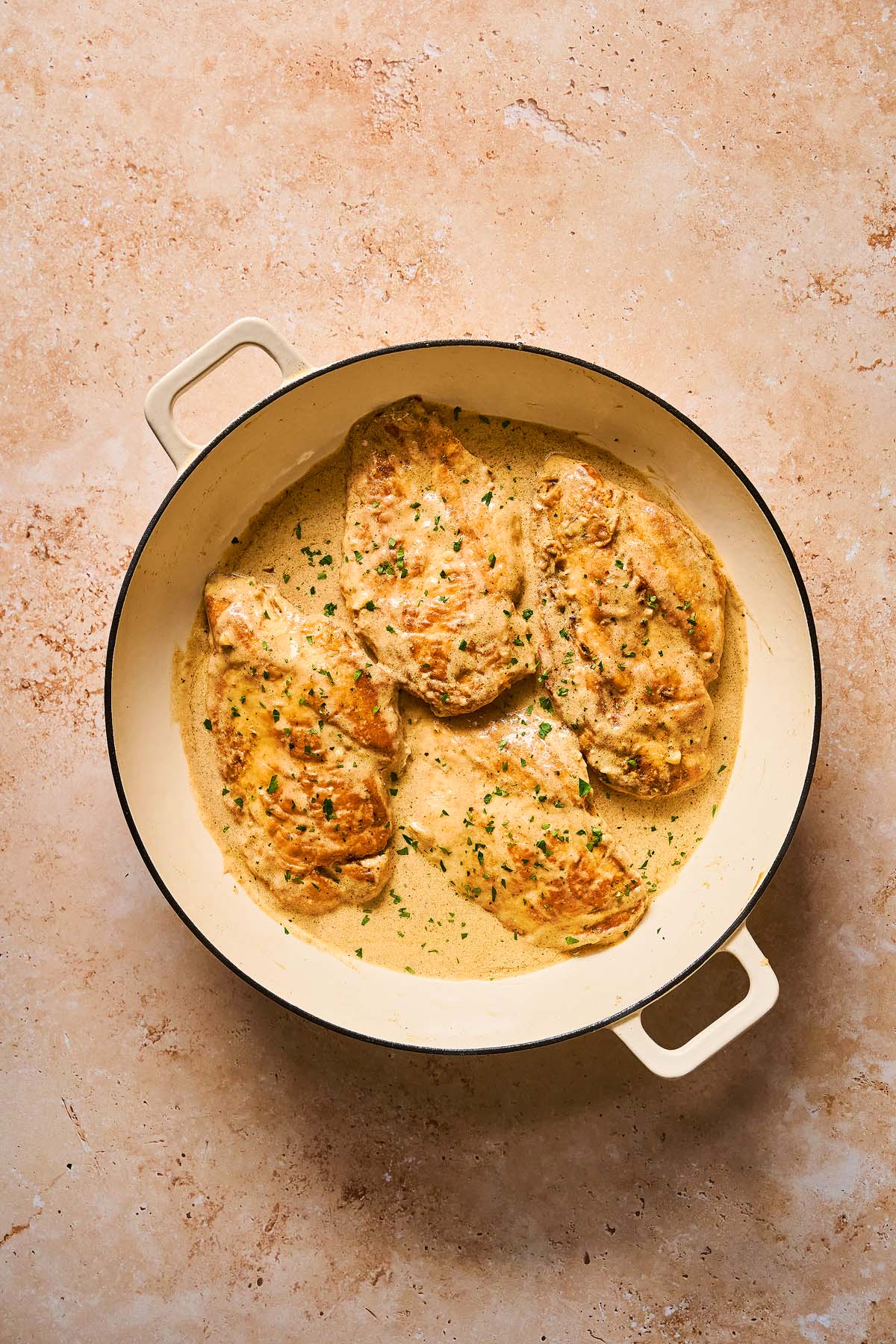 Creamy Chicken Limone breasts garnished with fresh herbs in a round, cream-colored skillet on a tan surface.