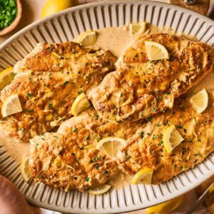 Hands holding a platter of creamy Chicken Limone breasts, garnished with lemon wedges and fresh herbs.