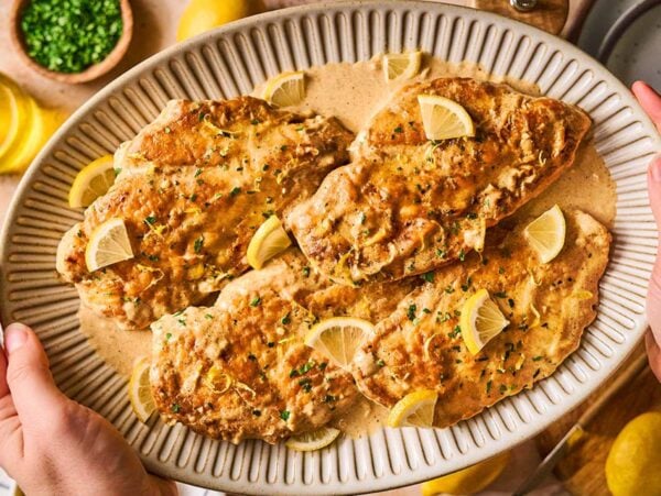 Hands holding a platter of creamy Chicken Limone breasts, garnished with lemon wedges and fresh herbs.