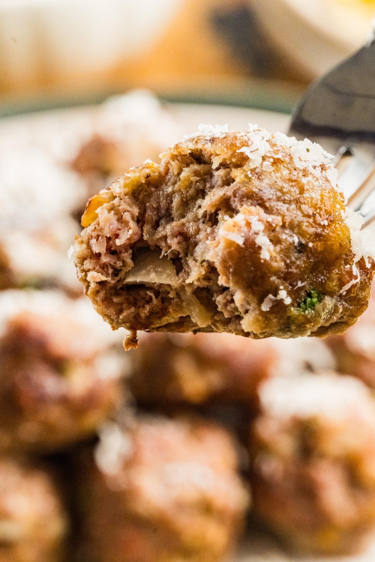 A close-up of a fork holding a meatball with a bite taken out, topped with grated cheese.