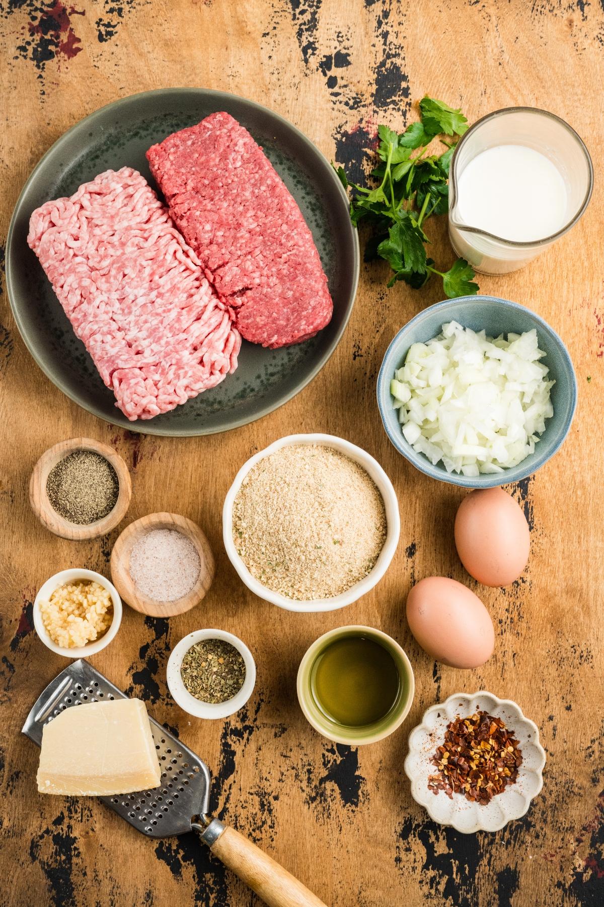 Top-down view of raw ground meats, chopped onion, eggs, spices, milk, herbs, oil, cheese, and breadcrumbs.