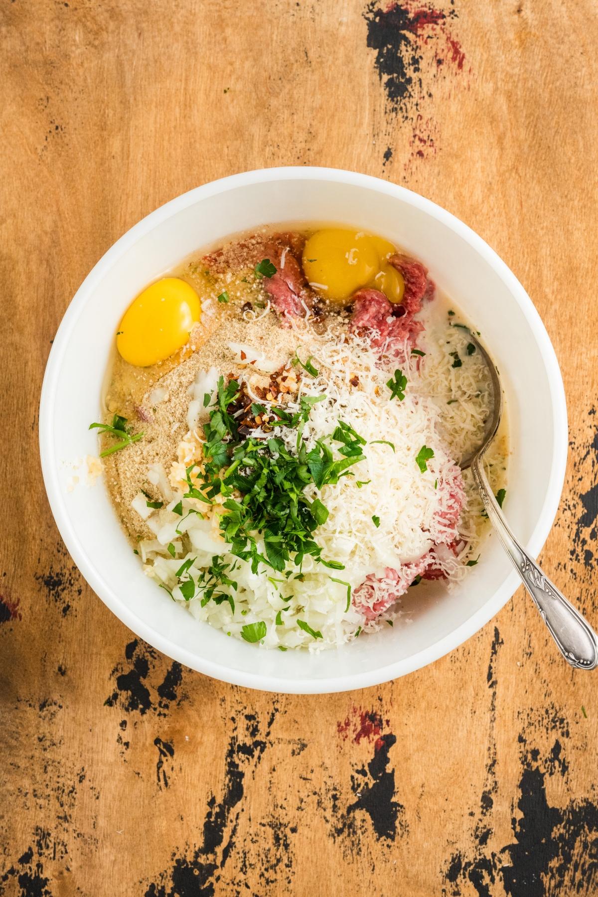 A bowl with ground meat, eggs, chopped onion, grated cheese, parsley, and breadcrumbs on a wooden surface.