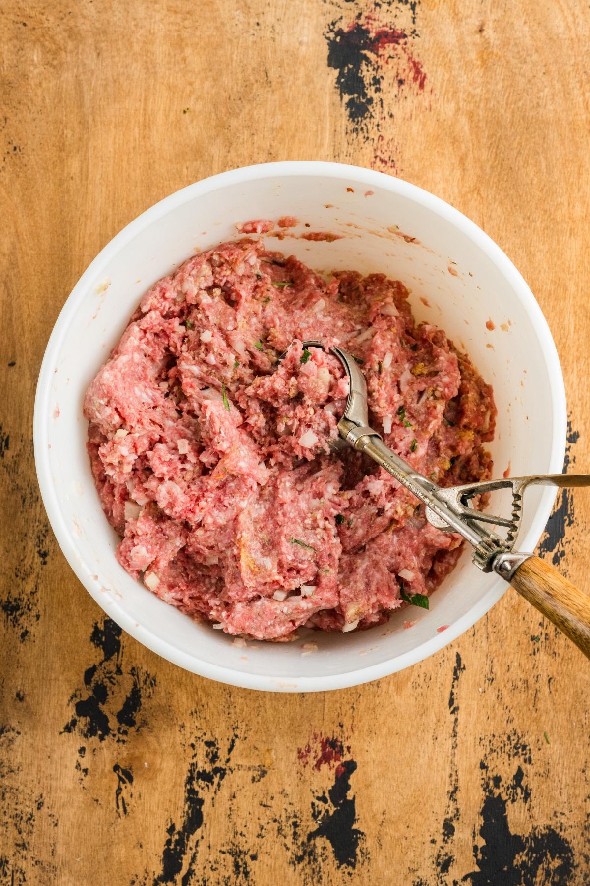A white bowl of raw ground meat mixture with a metal scoop on a wooden surface.