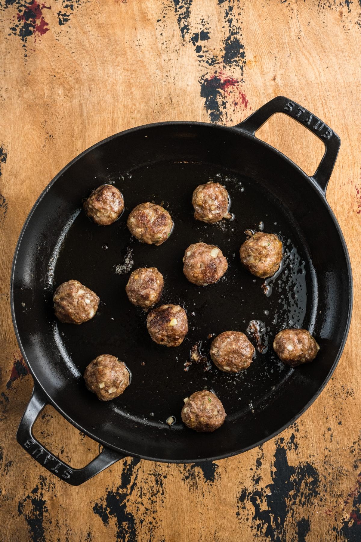 Meatballs cooking in a black skillet on a wooden surface with dark stains.