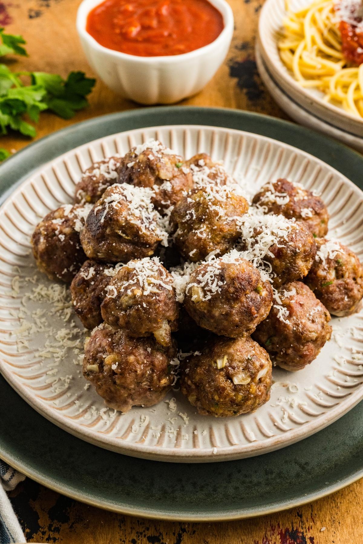 A plate of meatballs topped with grated cheese, with marinara sauce and spaghetti in the background.