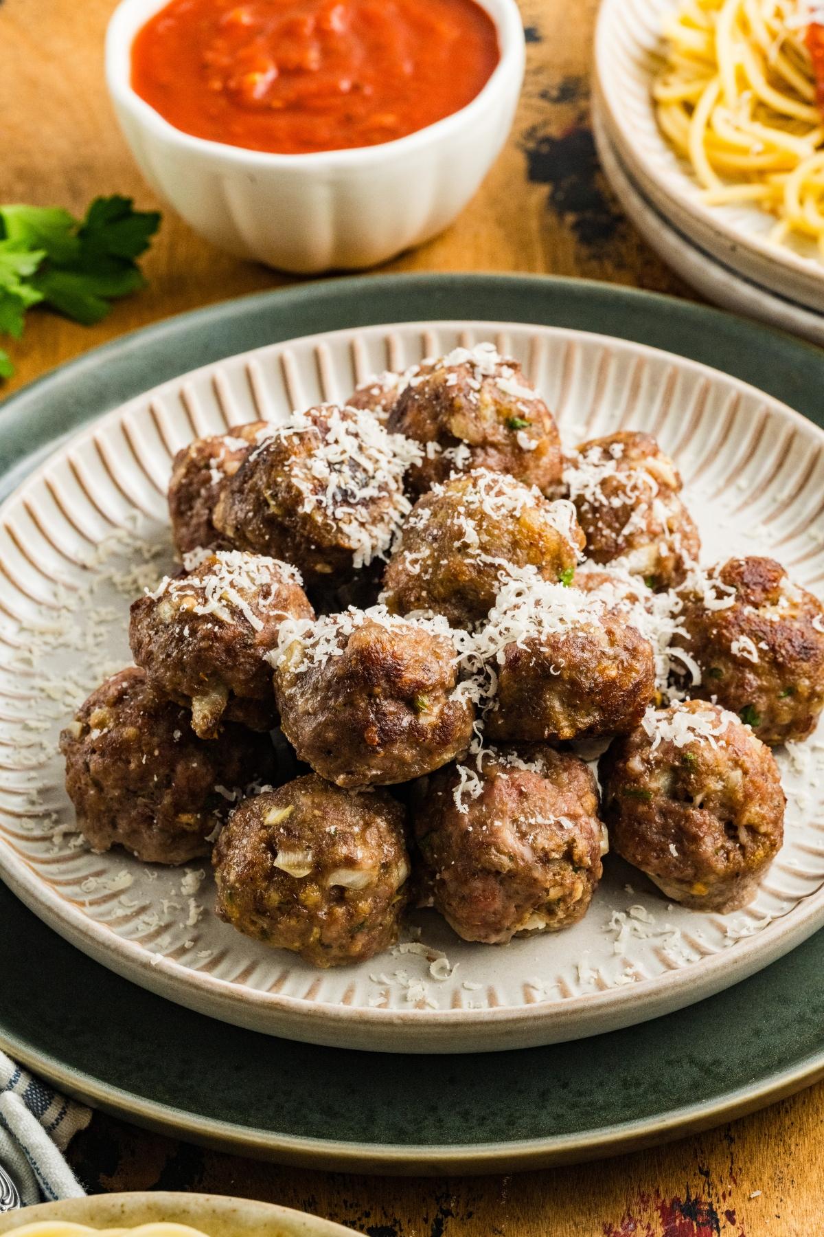 A plate of meatballs topped with grated cheese, with marinara sauce and spaghetti in the background.