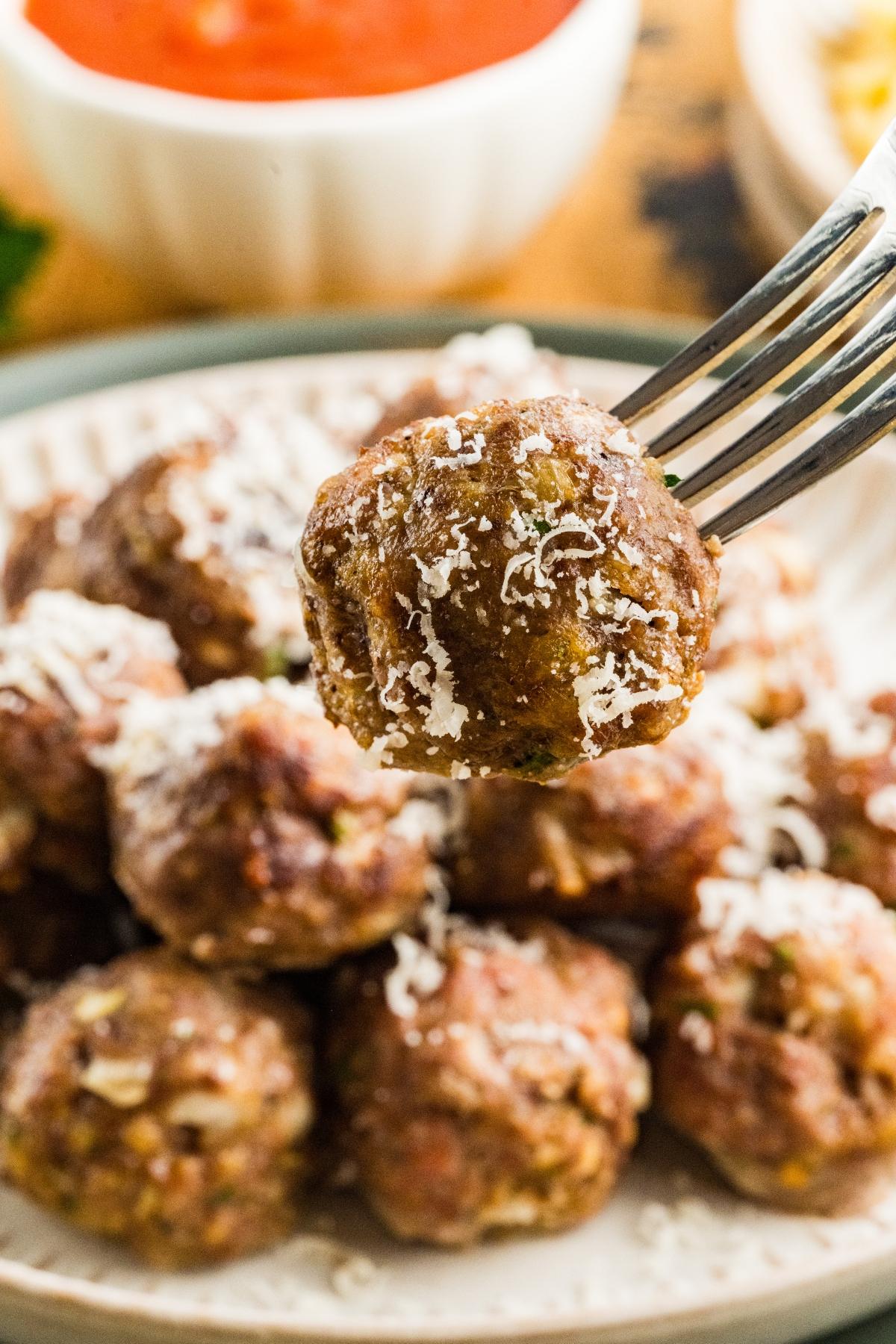 A fork holding a meatball topped with grated cheese, with more meatballs and sauce in the background.