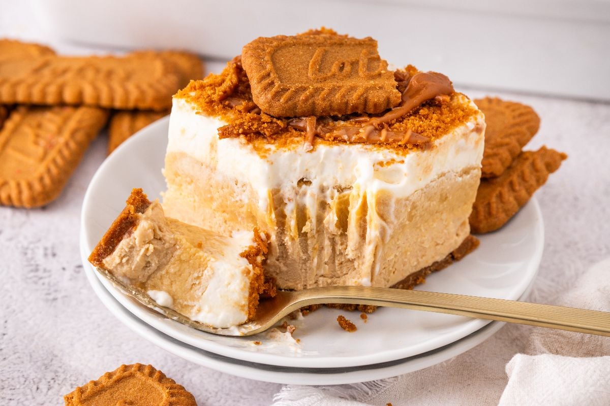 A creamy, layered Biscoff lush dessert topped with crumbled cookies and a whole cookie, served on a white plate with a fork.