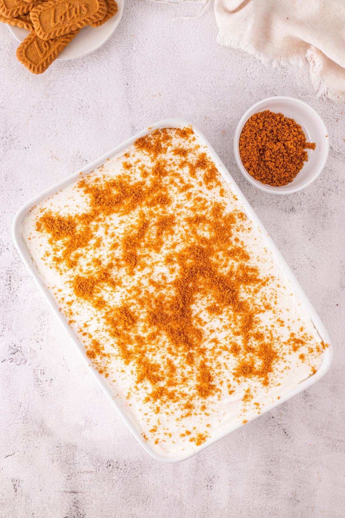 Rectangular dish of creamy dessert topped with crushed cookies, next to a bowl and whole cookies on a light surface.