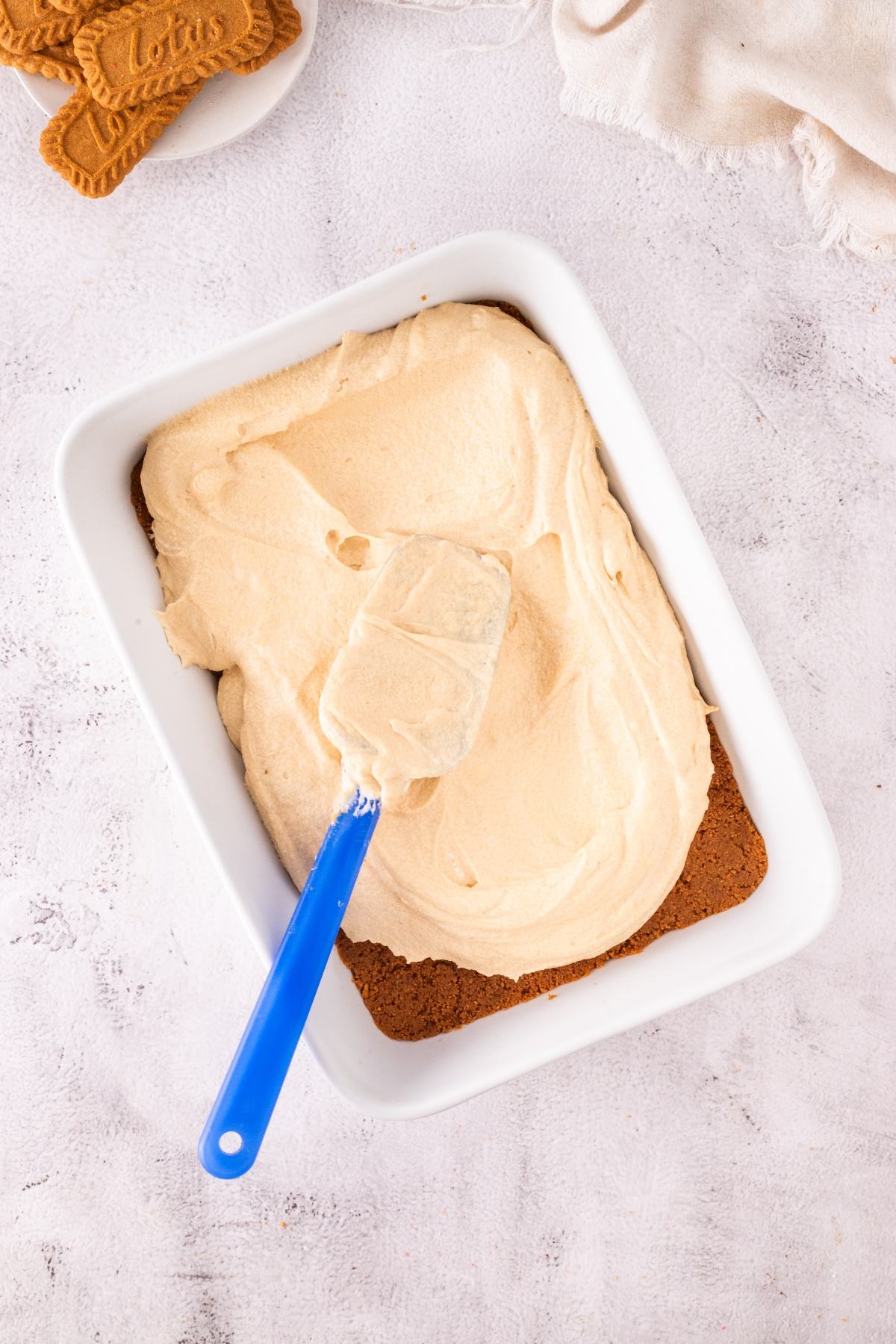 A blue spatula spreads creamy frosting over a dessert base in a white rectangular dish.