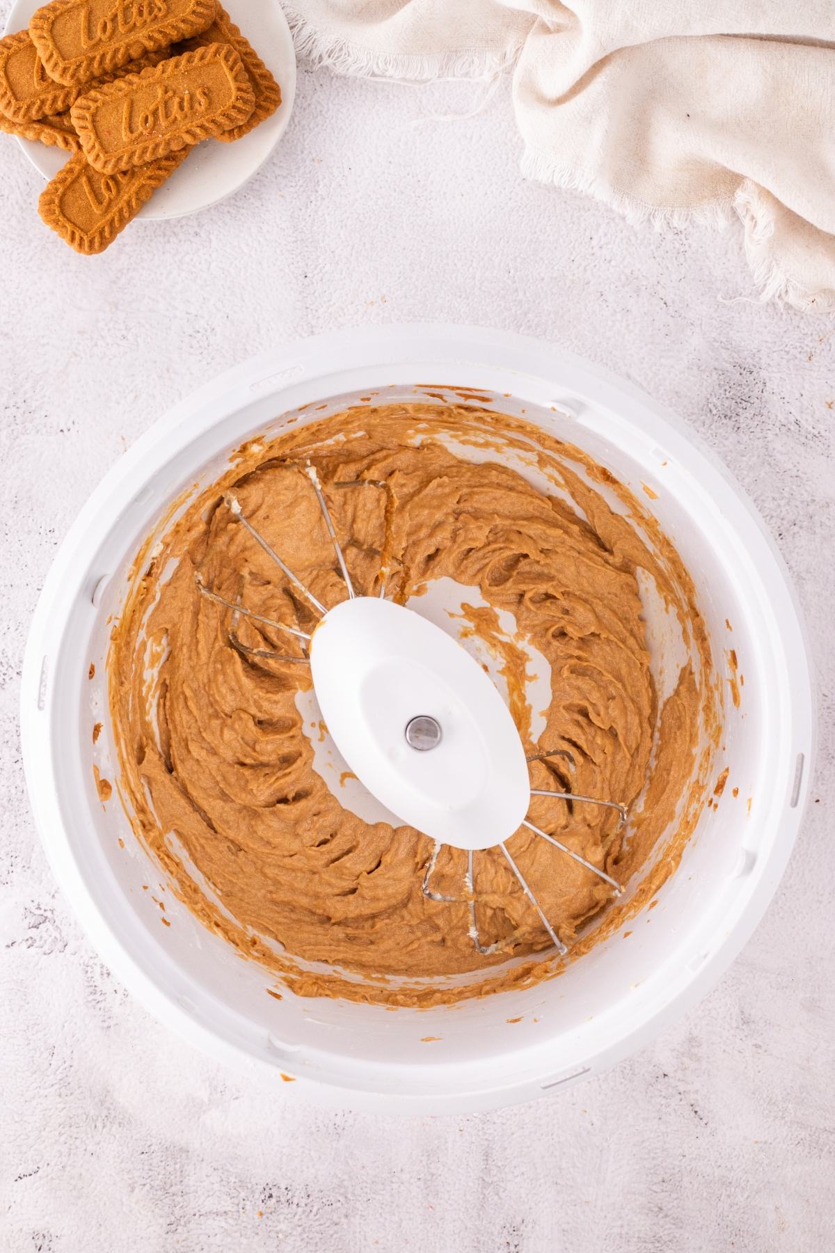 A mixing bowl with creamy brown batter and beaters, next to a plate of Biscoff cookies on a white surface.